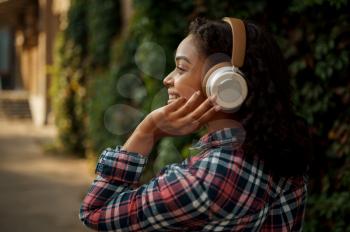 Cheerful woman in headphones listening to music in summer park. Female music fan walking outdoors, girl in earphones, green bushes on background