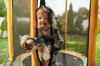 Kid holds funny dog on trampoline in the garden, best friends. Child with puppy leisures on backyard. Little girl and her pet having fun on playground outdoors, happy childhood