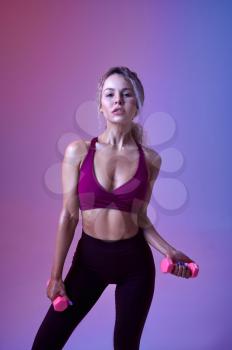 Young slim woman with dumbbells poses in studio, neon background. Sportswoman at the photo shoot, sport concept, active lifestyle motivation