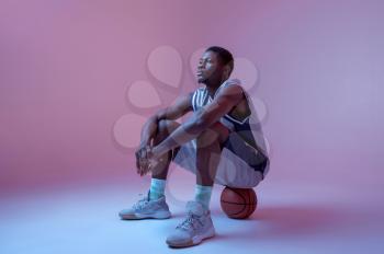 Basketball player sitting on ball in studio, neon background. Professional male baller in sportswear playing sport game, tall sportsman