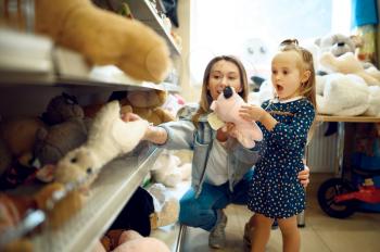 Mom and pretty little girl choosing soft toys in kid's shop. Mother and adorable daughter near the showcase in toyshop, happy childhood, family makes a purchase in store
