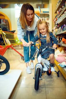 Mom and pretty little girl choosing bicycle in kid's store. Mother and adorable daughter near the showcase in toyshop, happy childhood, family makes a purchase in shop