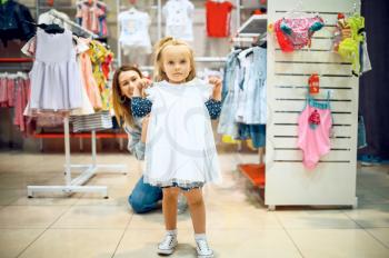 Mother and little baby trying on dress in kid's store. Mom and adorable girl near the showcase in children's shop, happy childhood, family makes a purchase in market