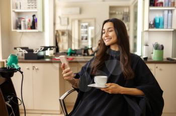 Smiling female customer with phone and cup of coffee in hairdressing salon.Woman sitting in chair in hairsalon. Beauty and fashion business, professional service