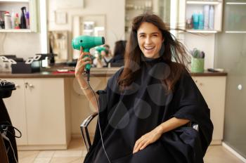 Happy female customer using hairdryer in hairdressing salon.Woman sitting in chair in hairsalon. Beauty and fashion business, professional service