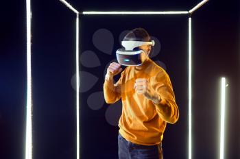 Young gamer fighting in virtual reality headset and gamepad in luminous cube, front view. Dark playing club interior, spotlight on background, VR technology with 3D vision