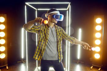 Man gaming in virtual reality headset and gamepad in luminous cube. Dark playing club interior, spotlight on background, VR technology with 3D vision