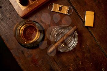 Cigar in ashtray and alcohol beverage in glass, lighter and guillotine on wooden table, top view, nobody. Tobacco smoking culture, specific flavor. Bad habits concept, smoker tools