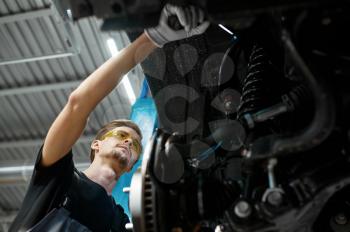 Male mechanic checks car suspension, auto service. Vehicle repairing garage, man in uniform, automobile station interior on background