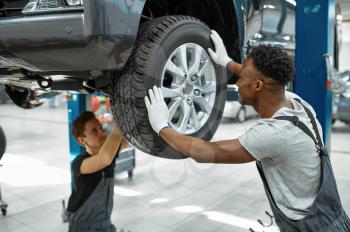 Two male workers fixes wheel, car service. Vehicle repairing garage, men in uniform, automobile station interior on background. Professional auto diagnostic