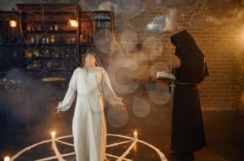 Male exorcist in black hood casting out demons from a woman. Exorcism, mystery paranormal ritual, dark religion, night horror, potions on shelf on background