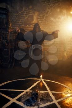 Male exorcist in black hood reads a spell near the magic circle. Exorcism, mystery paranormal ritual, dark religion, night horror, potions on shelf on background