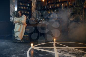 Young demonic woman sitting near magic circle with candles, demons casting out. Exorcism, mystery paranormal ritual, dark religion, night horror, potions on shelf on background