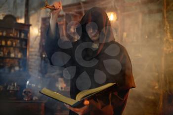 Male exorcist in black hood holds book of spells. Exorcism, mystery paranormal ritual, dark religion, night horror, potions on shelf on background
