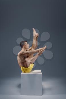 Male yoga sitting on the pedestal in studio, meditation position, grey background. Strong man doing yogi exercise, asana training, top concentration, healthy lifestyle