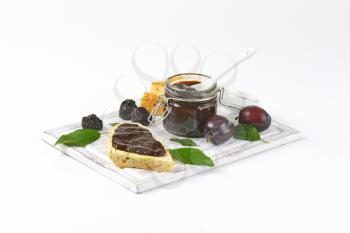 plum jam on a piece of baguette and in a jar, fresh plums and prunes on cutting board