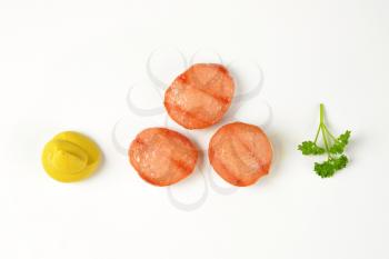slices of grilled sausage, mustard swirl and fresh parsley on white background