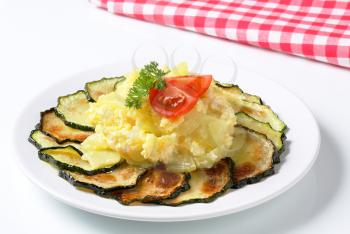 circle of thin slices of roasted zucchini with cooked potatoes, fried eggs, tomatoe and parsley on a white plate