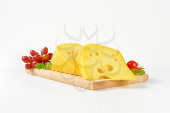 two wedges of Swiss cheese on wooden cutting board with red grapes next to it