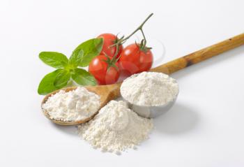 wheat flour on wooden spoon and in metal bowl, ripe tomatoes and fresh basil on white background