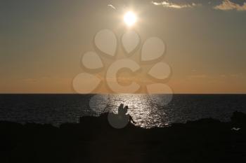 Silhouettes of two people sitting on rock on seashore at sunset
