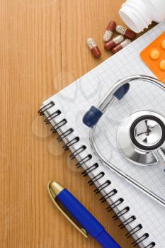 medical stethoscope with pills and notebook on wood