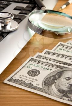 stethoscope at keyboard and dollars with pen on wooden table