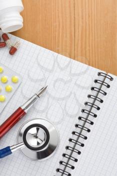 medical stethoscope with pills and notebook on wood