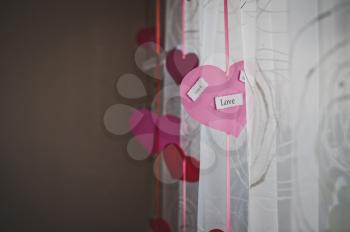 Pink paper hearts with inscriptions about love hung up on curtains.