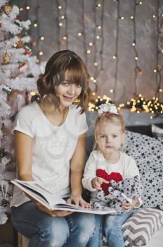 Mother and daughter looking in a beautiful book sitting around the Christmas decorated fir.