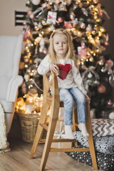 The child sits on a wooden chair beside the Christmas tree.