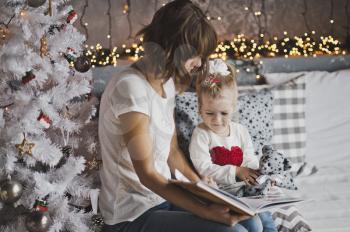 Little girl with mother reading a book sitting under the Christmas tree.