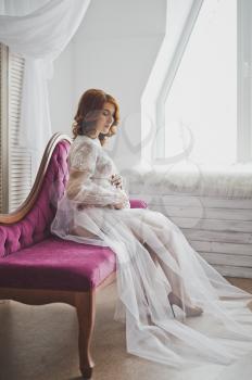 Pregnant girl in a semitransparent negligee sitting on the sofa.