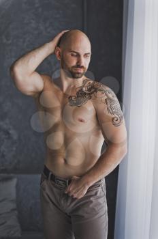 Portrait of a young bodybuilder with tattoos standing by the window.