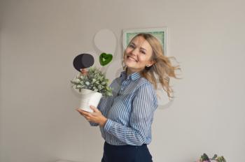 The girl in front of a white wall holding a pot of flowers and a sign.