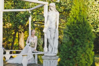 Bride in white dress sits on a bench in the garden.