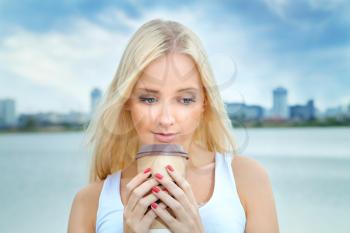 young blonde woman drinking coffee on a summer morning by the river with views of the city. Lifestyle