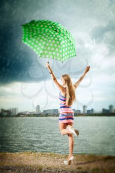 happy girl under an umbrella walks in the rain during the summer along the river against the background of the city