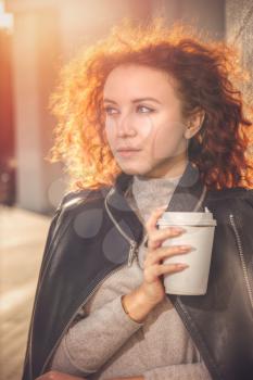 red-haired girl with a glass of coffee is standing by the wall. Portrait at sunset