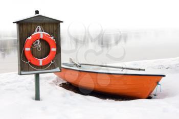 safety and lifesaving concept: orange lifeboat and orange lifebuoy on a snow-covered riverside in winter