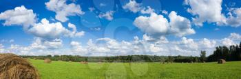 Panorama of field with haystacks near the forest under blue sky with white clouds under sunlight