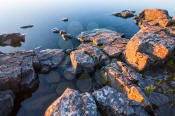 Rocks of stony coast in blue calm water of lake in yellow morning sun light in sunrise, minimalist nature landscape.