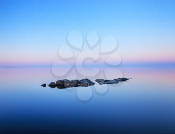 Tranquil minimalist landscape with rocks on the smooth surface of the lake with calm water with horizon with clear blue sky in twilight.