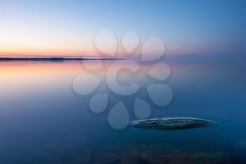 Tranquil minimalist landscape with rock in calm water of lake with smooth surface with horizon with clear blue sky in twilight, simple beautiful calm natural blue background.