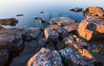 Rocks of stony coast in blue calm water of lake in yellow morning sunlight in sunrise, minimalist nature landscape
