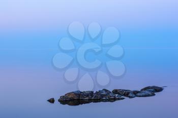 Tranquil minimalist landscape with rocks on the smooth surface of the lake with calm water with horizon with clear blue sky in twilight, simple natural blue background