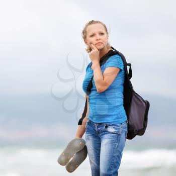 Pretty young blonde woman in a blue t-shirt and jeans with a backpack standing on a sea background.  Shallow depth of field. Focus on model.