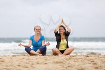 Two beautiful smiling young woman sitting in lotus pose on the sand on the beach and relax. Shallow depth of field. Focus on the models.