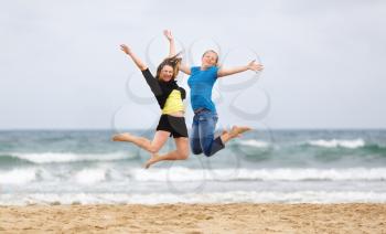 Two young smiling women jumping on the beach against the sea and cloudless sky. Shallow depth of field. Focus on the models.