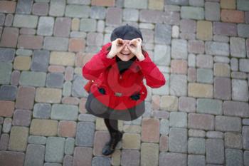 Photo of a smiling woman in a red jacket and a gray beret with hands that did the eyes like binoculars. The model stands on the sidewalk tile and looks up at the camera. Top view. Shallow depth of fie
